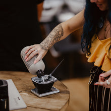 Person pouring a colour product from a bottle into a small container on a wooden table.