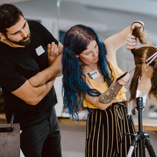 Two people working on a hair styling session with a third person observing.